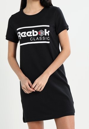 Kobieta ubrana w czarną sukienkę z krótkim rękawem z logo "Reebok Classic" i czerwonym emblematem gwiazdy na piersi, ręce splecione z przodu.