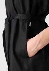 Μαύρο λινό ρούχο με δέσιμο στη μέση, πλαϊνή τσέπη και μικρή ετικέτα της μάρκας. Απαλή υφή με χαλαρή, ροή σχεδίαση.