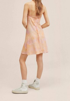 Roze tie-dye jurk met een fit-and-flare silhouet, verstelbare banden en een knoopdetail op de rug, in combinatie met witte enkelschoenen.