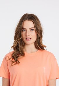 Korte mouwen, koraal T-shirt van zachte katoen. Voorzien van een kleine witte palmboomafbeelding op de linkerborst. Relaxte pasvorm met ronde hals.