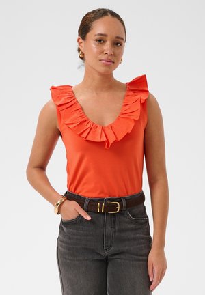 Femme aux cheveux bouclés portant un haut sans manches orange à volants, un jean foncé, des boucles d'oreilles créoles dorées, un bracelet et une ceinture marron avec une boucle dorée.