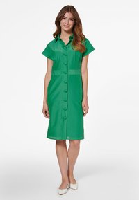 Grünes Hemdkleid mit Kragen, kurzen Ärmeln und taillierter Silhouette. Glatte Textur und knielanges Design. Weiße Schuhe.