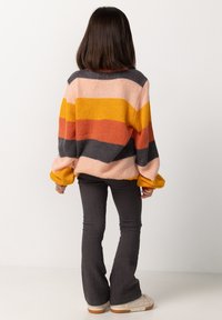 Pull surdimensionné avec des rayures horizontales en rose, orange, gris et jaune. Pantalons évasés côtelés gris foncé, chaussures moelleuses beige.