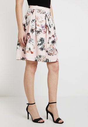 Frau trägt einen knielangen, blassrosa Rock mit großen Blumenmustern und schwarze, hochhackige Sandalen mit Knöchelriemen.