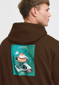 Brun hoodie med et rektangulært grafisk billede af en dessert på en sølvfarvet tallerken, med et kirsebær og et lille hvidt flag, blå-grønt baggrund.