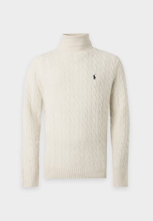 Jeune homme debout portant un pull à col roulé crème en maille torsadée, un pantalon cargo beige et des chaussures beiges slip-on sur un fond uni.