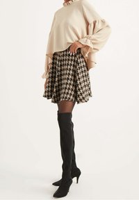 Béžový svetr s texturou a volány na rukávech, který je zkombinován s sukní v černobílém houndstooth vzoru. Modelka m�á na sobě černé vysoké kozačky na jehlovém podpatku.
