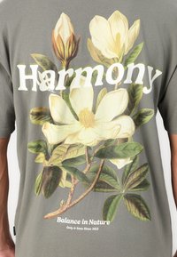 Grå bomulds t-shirt med et stort blomstermotiv, der viser hvide og gule magnolia-blomster og teksten "Harmoni." Nedenfor står der "Balance i Naturen."