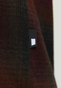 Primo piano di tessuto di lana marrone scuro e nero con un'etichetta per la cura nera piccola che presenta un simbolo rettangolare bianco.