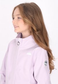 Chaqueta morada clara hecha de material suave, con un alto cuello, cierre de cremallera y pequeño logotipo de paraguas en el pecho con un parche circular en la manga.