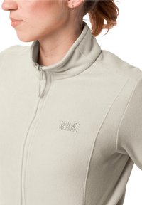 Ljusbeige fleecejacka med hög krage, dragkedjestängning och broderad logotyp på vänster bröst. Mjuk textur och minimalistisk design.