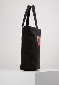 Svart tote-väska med långa, svarta tygremmar och en röd logotyp på framsidan, med rektangulär form och slät yta.
