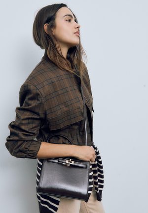 Giacca cropped a quadri marrone, borsa in pelle nera con un gancio argentato, tenuta da una modella su uno sfondo chiaro. Accenti a righe visibili.