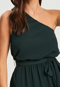 Sukienka na jedno ramię w ciemnozielonym kolorze, wykonana z teksturowanej tkaniny, z wiązanym detalem w okolicy talii i delikatnymi fałdami dla swobody ruchu.
