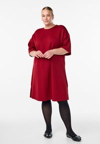Červené voľné šaty s krátkymi rukávmi, vyrobené z mäkkého materiálu. Majú bočné rozparky a okrúhly výstrih, kombinované s čiernymi pančuchami a plochými topánkami.