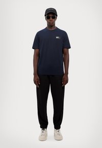 Navy t-shirt with a small logo, black joggers with ankle cuffs, and white sneakers. Accessories include a black cap and sunglasses.