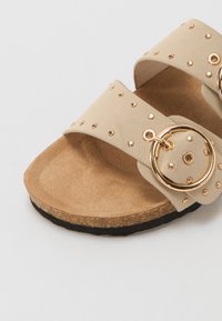 Sandales beiges à double bride, ornées de cercles décoratifs en or et de clous, avec une semelle intérieure texturée et une semelle en liège.