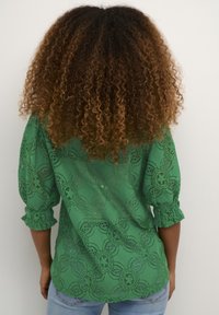 Zelena čipkasta bluzica s 3/4 rukavima, koja ima cvjetne uzorke i dizajn skupljenih manžeta, nosi se s svijetloplavim farmerkama. Pogled s leđa.