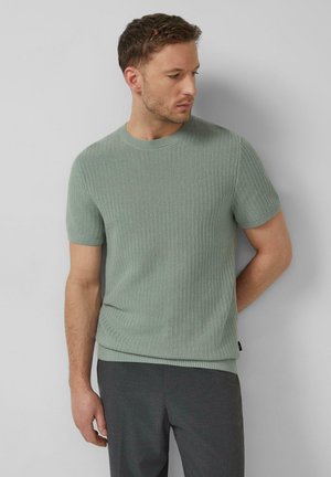 Man in een saliegroene, korte mouw, gebreide trui en donkergrijze broek staat tegen een lichtgrijze muur en kijkt naar beneden naar de zijkant.