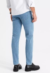 Muž v světle modrých džínsech, černých kožených polobotkách a bílé košili s dlouhým rukávem, která má vyhrnuté rukávy, stojí na bílém pozadí.