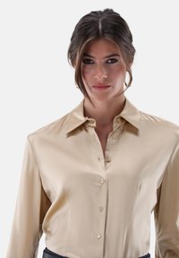 Goldene Satinbluse mit langen Ärmeln, spitzem Kragen und Knopfverschluss vorne. Verfügt über zarte Knöpfe und eine glatte Textur.