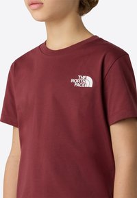 Vinrød bomulds-t-shirt med et hvidt logografik på øverste venstre side, rund halsudskæring og korte ærmer. Blød tekstur.