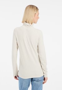 Haut col roulé à côtes, beige clair, avec des manches longues et un ourlet légèrement arrondi. Texture douce, branding minimal sur le côté.
