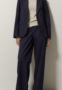Blazer tailored di un blu navy con un bottone unico, abbinato a pantaloni a gamba larga coordinati. Il blazer ha una vestibilità strutturata e una texture liscia.