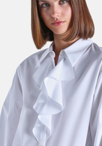 Jeune femme aux cheveux bruns et raides portant une chemise blanche avec un grand volant sur le devant, sur un fond clair.