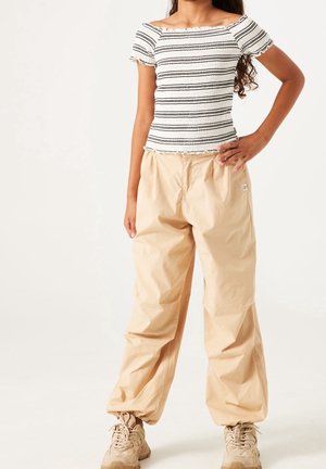 Jeune personne portant un haut blanc et noir rayé, épaules dénudées, avec un pantalon beige ample et des baskets beiges, debout avec une main sur la hanche.
