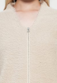 Lichtbeige ritscardigan gemaakt van een zachte textuurstof met een fuzzy afwerking, met een zilveren rits en een sjawlboord.