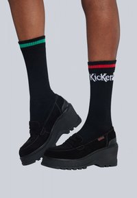 Mocasines de plataforma negros hechos de ante, con costuras decorativas, combinados con calcetines negros acanalados con franjas verdes y rojas.