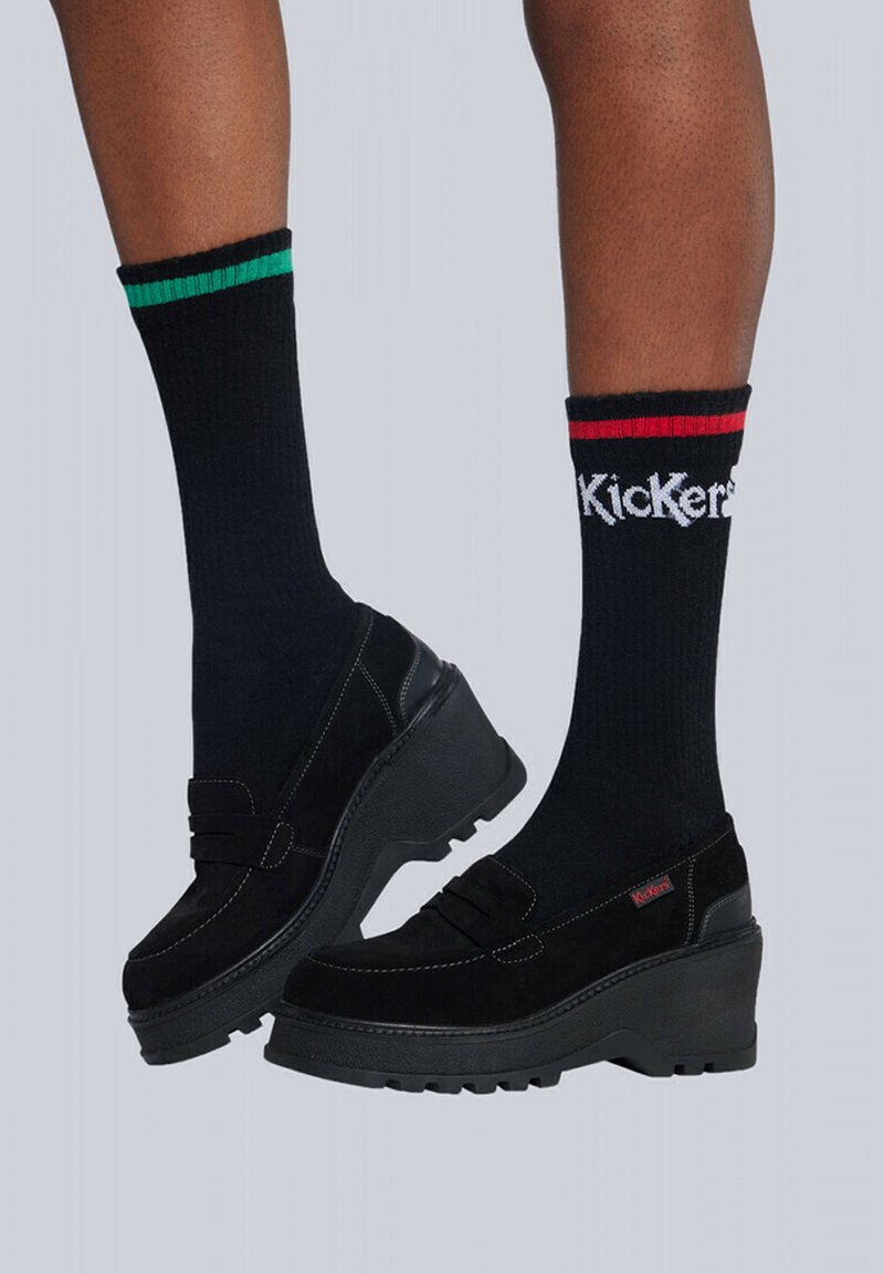 Mocasines de plataforma negros hechos de ante, con costuras decorativas, combinados con calcetines negros acanalados con franjas verdes y rojas.