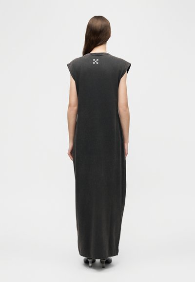 Γυναίκα με μακριά καστανά μαλλιά, φορώντας αμάνικο μαύρο φόρεμα μέχρι τον αστράγαλο με λευκό λογότυπο βέλους στην πάνω πλάτη, όρθια με την πλάτη γυρισμένη.