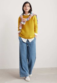 Gul ribbad tröja, vit skjorta med krage under, i kombination med vida blå denimbyxor. Mångfärgad mönstrad scarf hängande över axlarna.