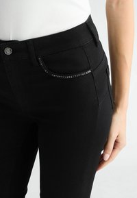 Un jean en denim noir présente une coupe slim, un bouton argenté et un accent de perles scintillantes le long de la poche avant. Le matériau est lisse avec une légère élasticité.