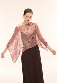 Femme aux cheveux tirés en arrière, portant de grandes boucles d'oreilles en or, un châle rose transparent à motifs floraux sur une robe noire, debout les bras légèrement étendus.