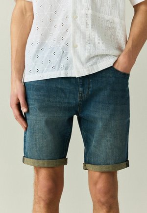 Mann trägt blaue Jeansshorts mit Umschlag und ein weißes kurzärmliges Hemd mit Knöpfen und Ösenmustern, eine Hand in der Hosentasche.