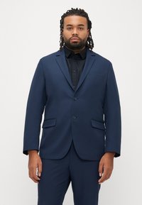 Marineblauw pak met een gestructureerd jasje, enkelrijig ontwerp, revers met inkeping, twee knopen en zijzakken, gecombineerd met een zwart overhemd.