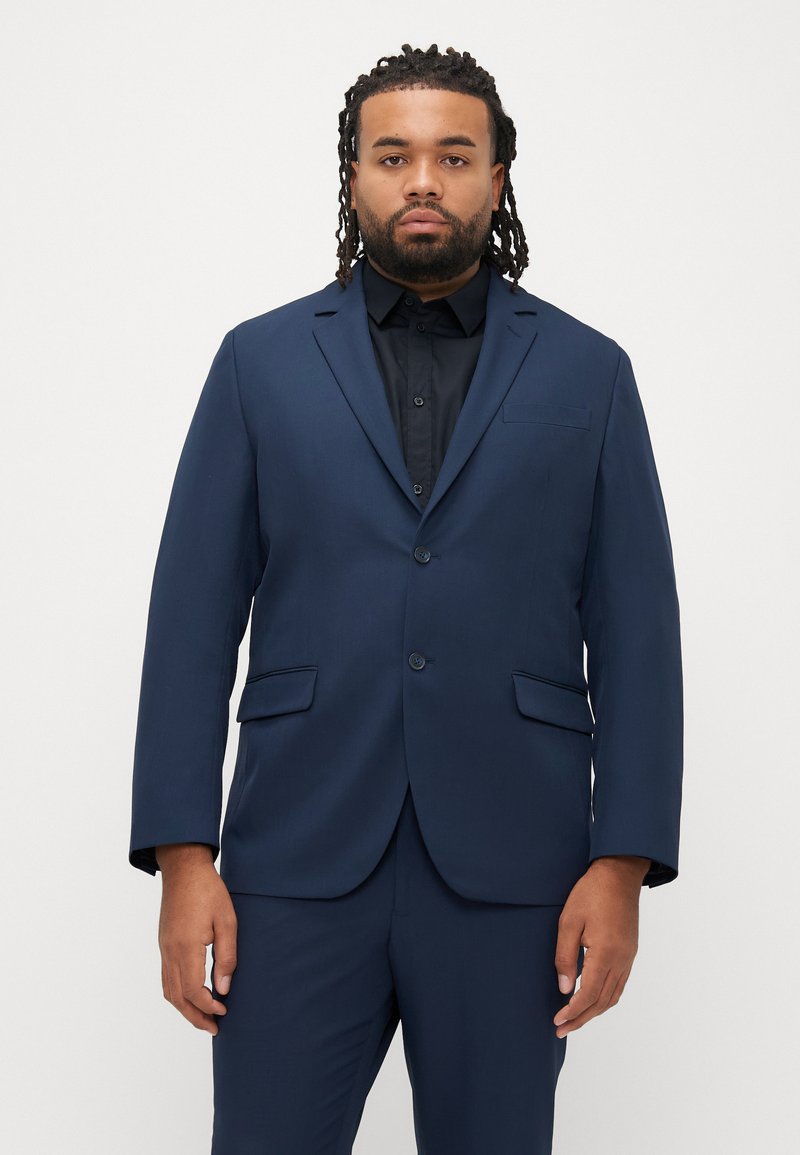 Marineblauw pak met een gestructureerd jasje, enkelrijig ontwerp, revers met inkeping, twee knopen en zijzakken, gecombineerd met een zwart overhemd.