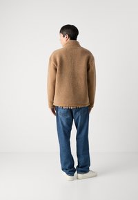 Brun fleecejakke med høj krave, tekstureret overflade og drop-skulder, parret med blå jeans og hvide sneakers.