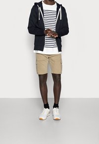 Námořnická mikina na zip, bílo-námořnicky pruhované tričko, khaki cargo šortky, černé ponožky a bílé tenisky s oranžovými detaily.