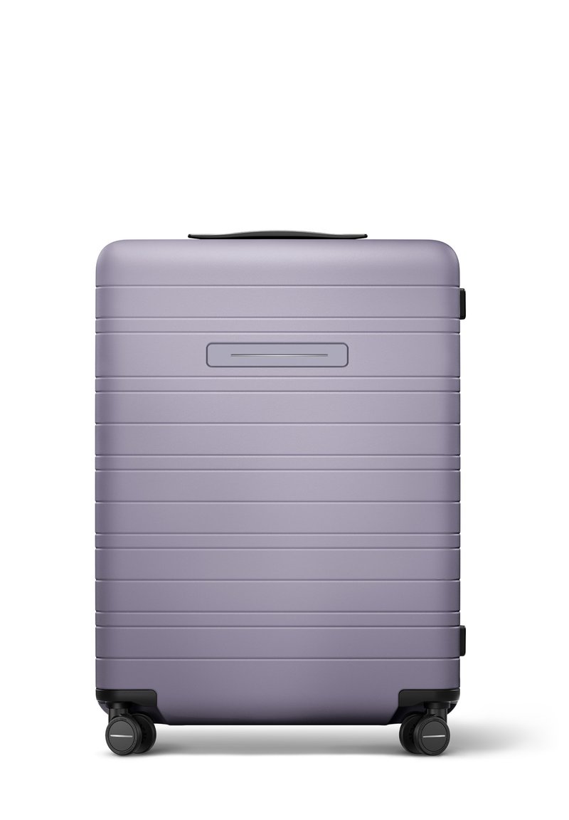 Valise rigide de couleur violet clair avec une finition mate, présentant des rainures horizontales, une poignée supérieure et quatre roulettes noires.