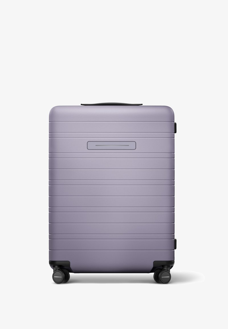Valise rigide de couleur violet clair avec une finition mate, présentant des rainures horizontales, une poignée supérieure et quatre roulettes noires.