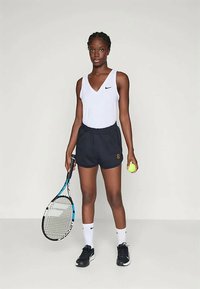 Αθλήτρια που στέκεται με ρακέτα τένις στο δεξί χέρι και μπάλα στο αριστερό, φορώντας λευκό αμάνικο μπλουζάκι, μαύρες βερμούδες, λευκές κάλτσες και μαύρα αθλητικά παπούτσια.