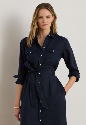 Žena v tmavomodrých šatách zapínaných na gombíky s vreckami na hrudi, vyhrnutými rukávmi a opaskom zviazaným v páse, stojaca s jednou rukou v vrecku.