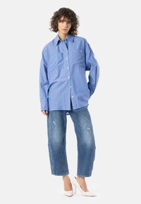 Blåstripete skjorte med knapper og to frontlommer, kombinert med slitte lyse denimjeans og kremfargede høye hæler.