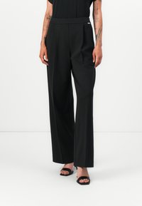 Pantaloni neri a gamba larga con una texture liscia, pieghe frontali sartoriali e una cintura comoda. Abbinati a tacchi aperti neri.