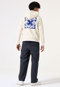 Felpa beige con una grafica a motivo foglia blu sulla schiena; abbinata a pantaloni cargo grigio scuro e sneakers bianche.