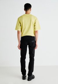 Geel T-shirt met korte mouwen, comfortabele pasvorm, gecombineerd met slim-fit zwarte jeans. De achterkant toont de onderkant van het shirt net boven de tailleband van de jeans. Zwarte schoenen.